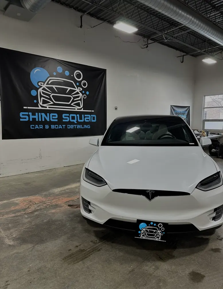 A white Tesla SUV parked in the Shine Squad shop in front of the Shine Squad car and boat detailing banner