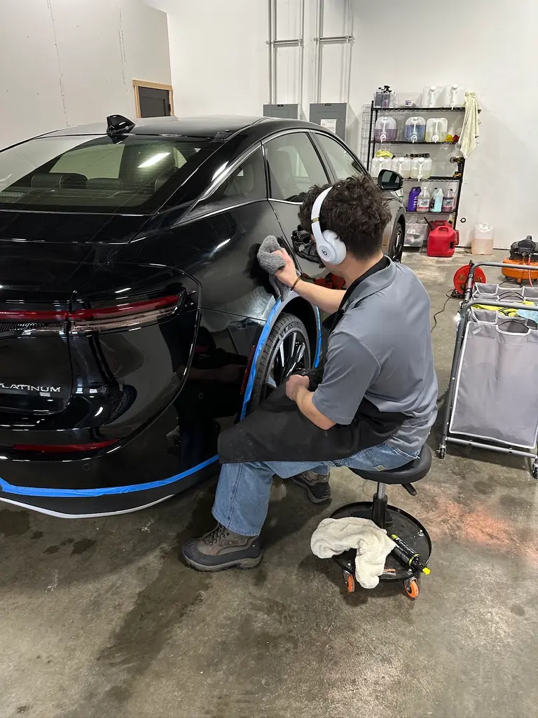 A Shine Squad technician hand-polishing the rear fender of a black sedan with blue tape protecting the trim