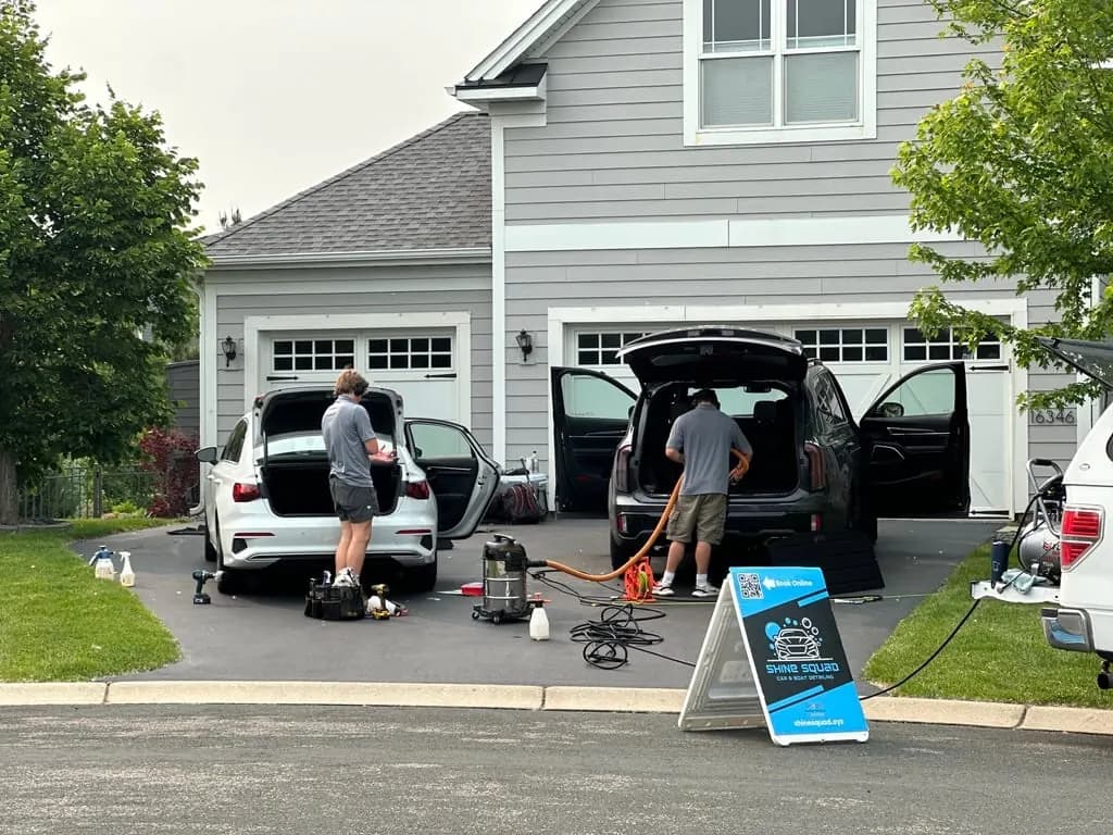 Two Shine Squad technicians detailing a pair of vehicles beside their branded A-frame sign